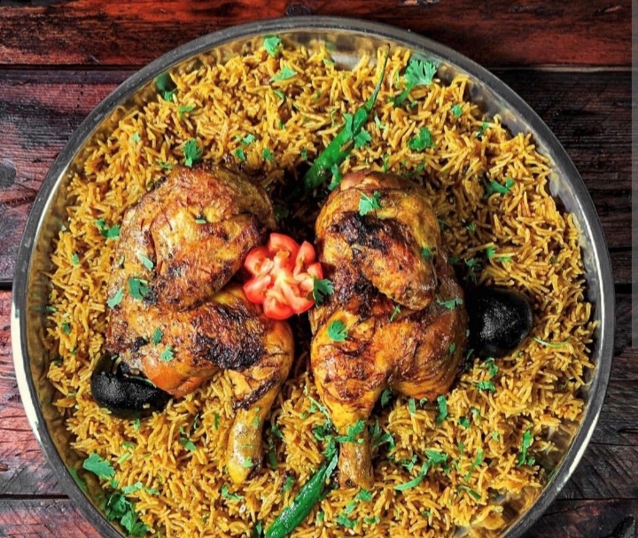 A serving platter with two pieces of grilled chicken resting on a bed of spiced rice, garnished with green chillies, coriander leaves, and a slice of tomato.